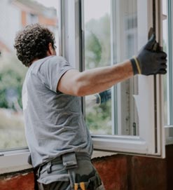 Person wearing a gray T-shirt, gloves, and a tool belt, seen from behind while adjusting the sash of an open white-framed casement window.