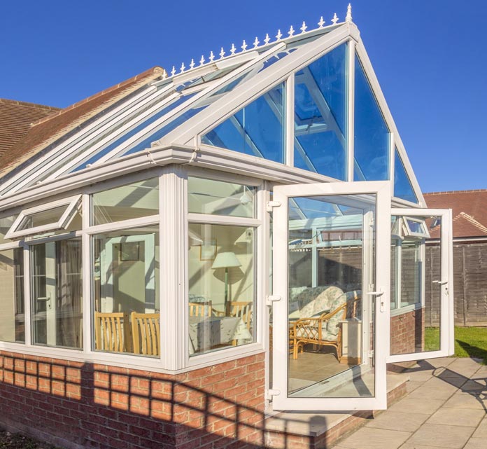 A bright glass conservatory attached to a brick house with white-framed windows and a steep triangular glass roof topped with decorative finials.