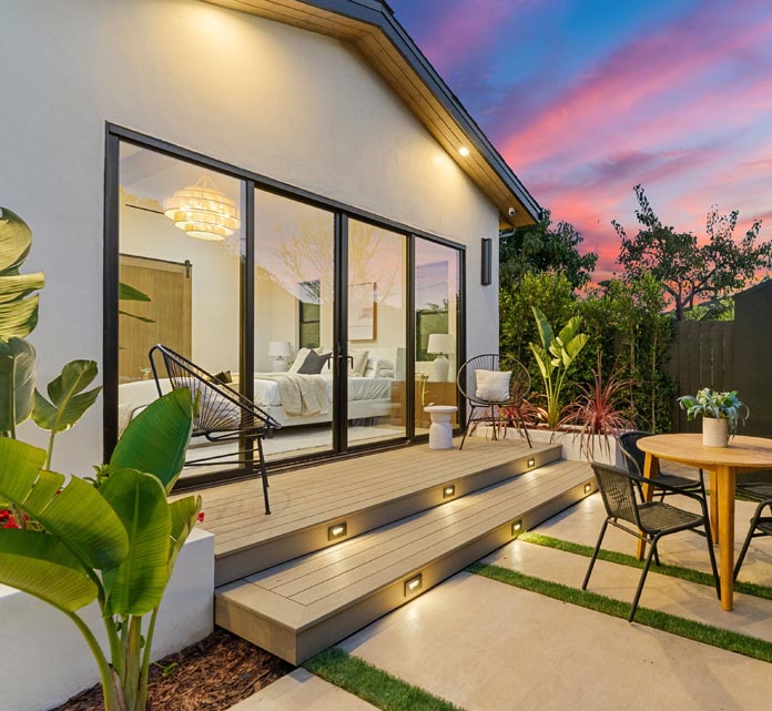 Modern cozy backyard patio at dusk with a small light-colored house extension featuring large black-framed sliding glass doors. A composite wood deck with recessed step lights leads to outdoor seating and a wooden dining table.