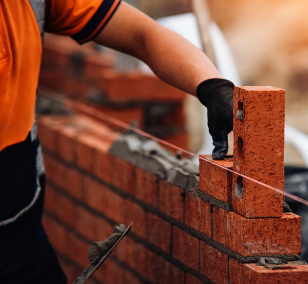 A close-up of a bricklayer’s hand wearing a black work glove, carefully placing a perforated red brick onto a bed of mortar while building a red-brick wall.