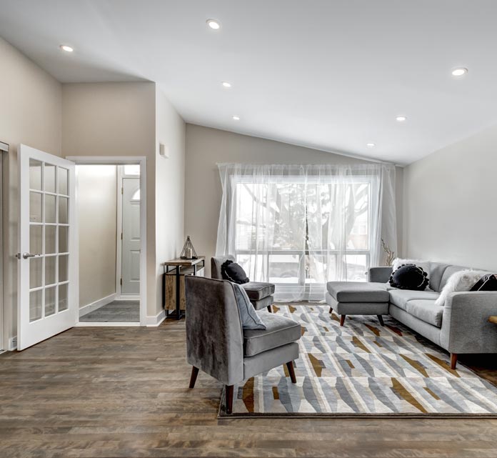 Bright, modern garage conversion, living room with a light gray sectional sofa and gray armchairs on warm wood flooring. A large window with sheer white curtains lets in natural light, the room has a sloped ceiling with recessed lights and minimal decor.