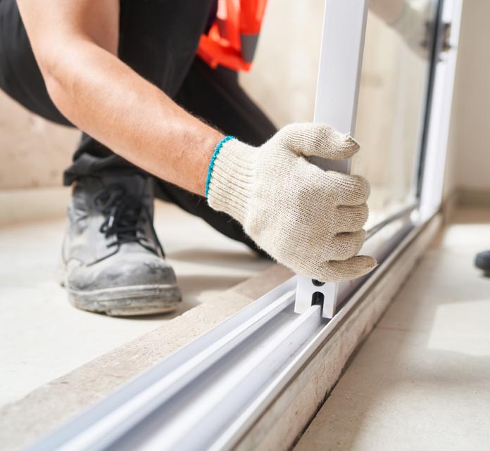 A close-up of a gloved hand adjusting a white sliding door set into an aluminum track on the floor. The person is kneeling, wearing work boot and a high-visibility vest, showing an installation in progress.