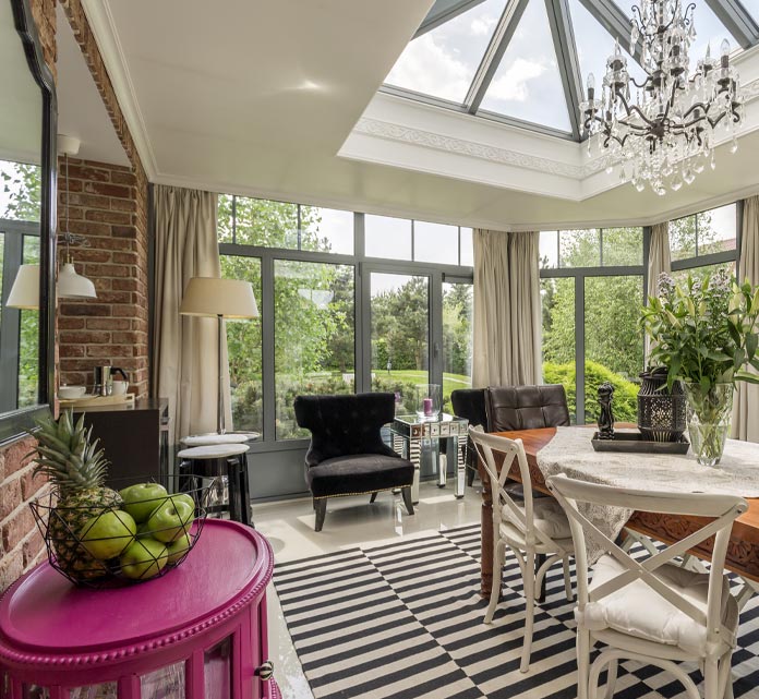 Bright orangery with large glass windows and Lantern Roof. The room has a wooden dining table with white cross-back chairs, a black-and-white striped rug, exposed red brick wall, pale tile floor, and neutral curtains framing views of a lush green garden.