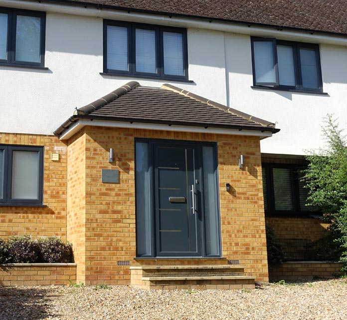 A modern house front entrance with a small brick porch topped by a tiled hipped roof. The dark grey front door has a vertical handle and narrow glazed sidelights. Three steps lead up from a gravel driveway to the porch.