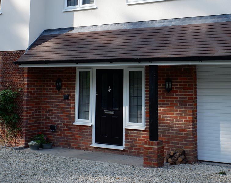 Front entrance of a suburban house featuring a central black door, flanked by narrow vertical leaded-glass sidelights. A small pitched porch roof with dark-brown tiles supported by a black post covers the doorway.