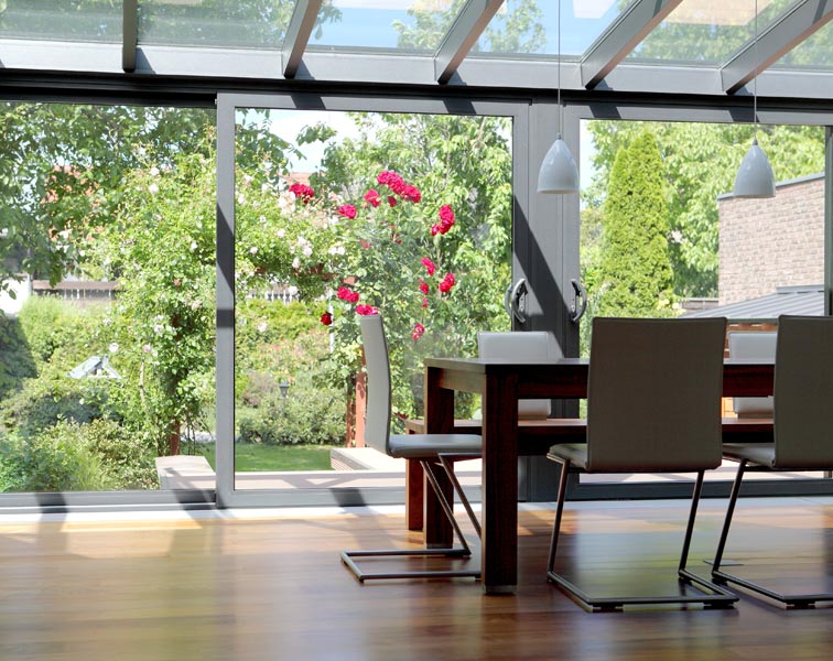 A bright, contemporary dining area in a modern aluminium conservatory with a slanted glass roof and large sliding glass doors opening to a green garden.