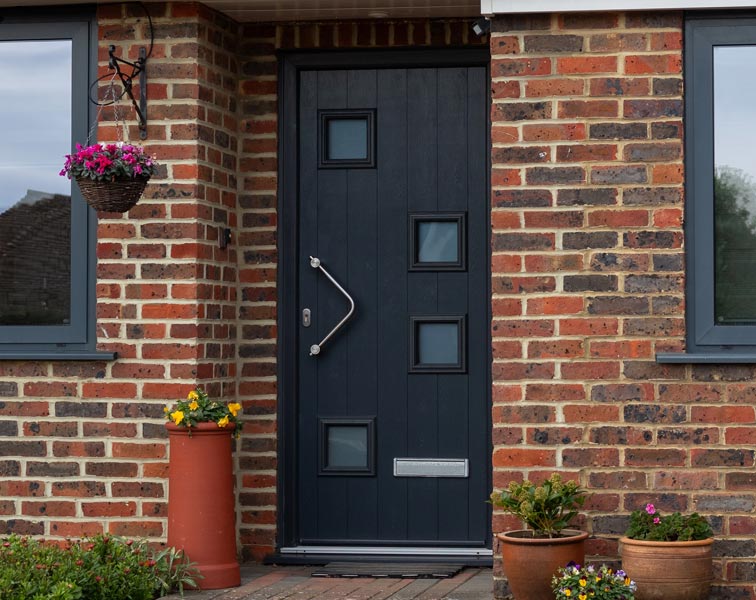 A modern dark grey front door with vertical paneling and four small square glazed windows arranged vertically, featuring a curved stainless-steel pull handle. The door is set in a red-brick exterior with gray-framed windows on either side.