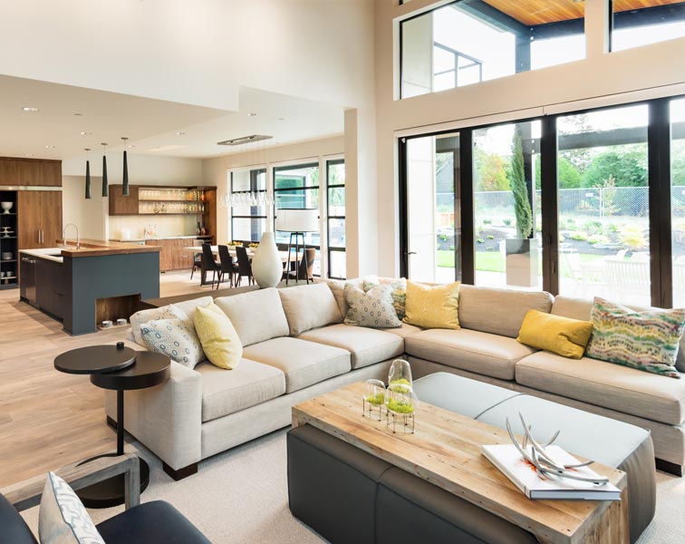 Bright open-plan living area with a large beige L-shaped sectional sofa adorned with patterned and yellow pillows. The room has floor-to-ceiling windows and sliding doors showing a landscaped yard, light wood flooring, high ceilings, and a modern kitchen with a dark island, pendant lights, and a dining table nearby.