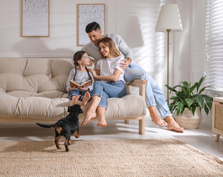 Warm, bright living room with a man, woman, and young girl—sitting closely together on a beige futon sofa. The girl holds an open book, while her parents smile and lean in. A small black-and-tan dog walks on a woven jute rug. Sunlight filters through blinds onto white paneled walls decorated with simple framed prints, creating a cozy, intimate atmosphere.