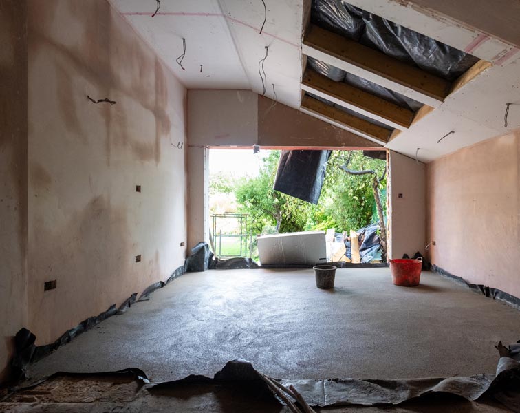Interior garage under renovation with freshly leveled concrete floor and newly plastered pale walls. The sloped ceiling shows exposed insulation and roof joists with hanging electrical wiring.
