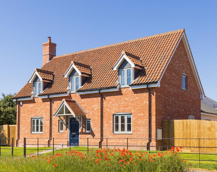 Well-kept two-story red brick house with steep terracotta-tiled roof, three dormer windows, and a chimney. The house has white-framed windows, a small gabled porch over a dark front door.
