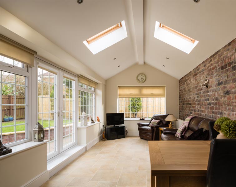 Bright orangery sunroom with vaulted ceiling and rectangular skylights. One wall is large glass doors and windows overlooking a fenced garden, opposite an exposed red brick wall. The room has beige tile flooring, dark brown leather armchairs with cushions, a side table with lamp, a wooden table with potted plant.