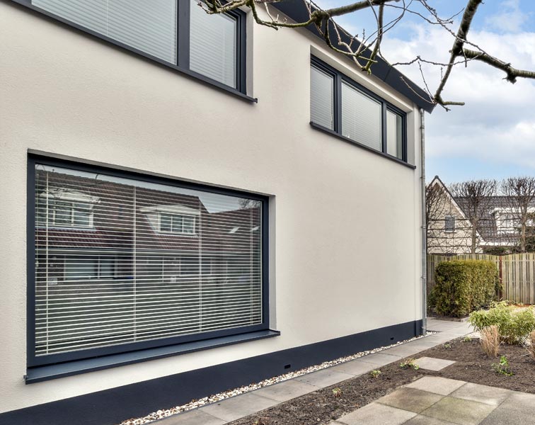 Modern two-story house exterior with smooth off-white rendered walls and dark gray trim. Large ground-floor window with horizontal blinds, two narrow upper-floor windows with closed blinds.