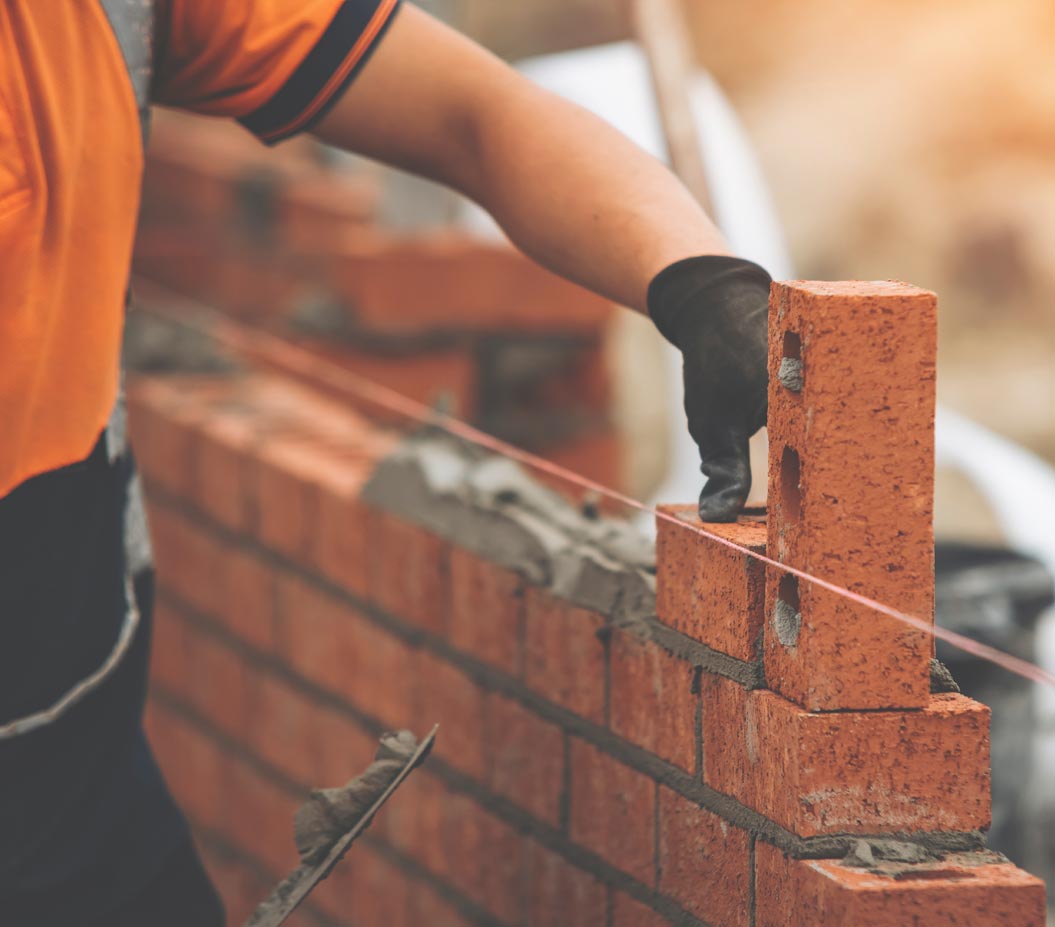A close-up of a bricklayer’s hand wearing a black work glove, carefully placing a perforated red brick onto a bed of mortar while building a red-brick wall.