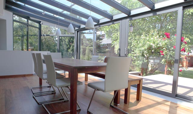 A bright, modern aluminium conservatory with a glass roof and large floor-to-ceiling sliding glass doors opening onto a lush garden.