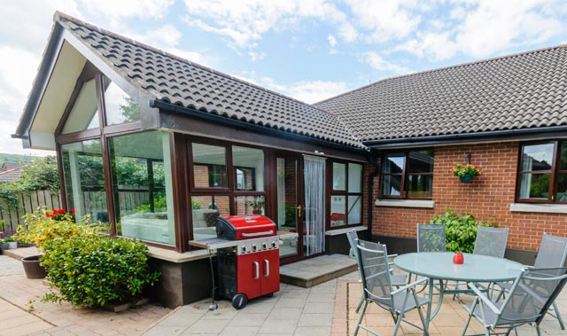 A sunny backyard patio beside a single-story brick house with a tiled sloped roof. A tiled roof conservatory with dark wooden frames and sliding glass doors extends from the house. 