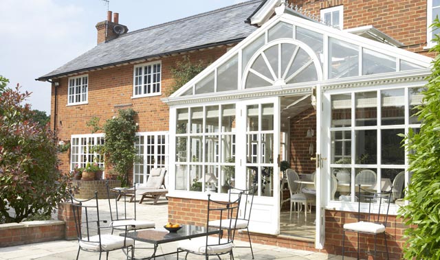 A sunny backyard showing the rear of a two-story red-brick house with a white-framed glass conservatory attached. The conservatory has a pitched glass roof with a decorative arched window at the gable.