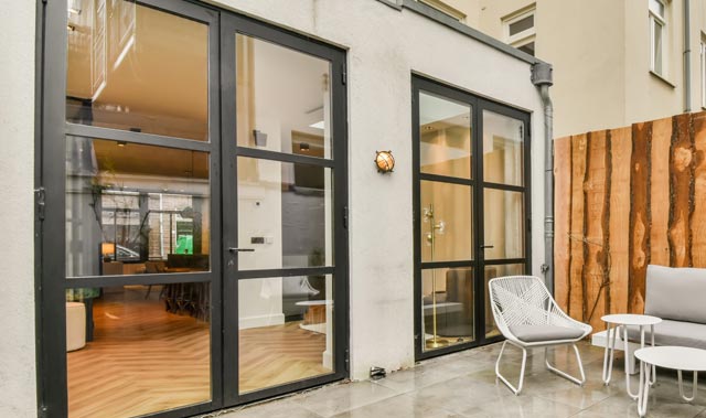 A small modern patio outside a ground-floor rear entrance with two large black aluminium framed glass heritage doors featuring a grid of horizontal and vertical mullions. Through the doors, a warm, well-lit interior with herringbone wood flooring can be seen.