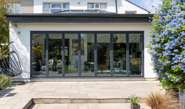 The rear exterior of a modern house featuring a modern extension and wide set of dark-framed glass bi-fold doors with six tall panels, opening onto a paved patio.