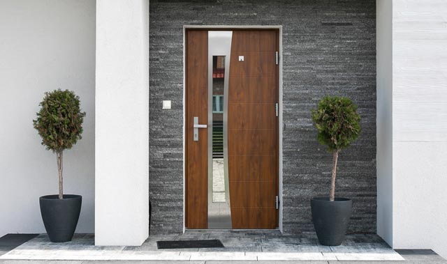 A modern, symmetrical front entrance with a composite  door featuring a narrow vertical glass panel offset to one side. The door is framed by a dark gray stacked-stone surround and has a horizontal metal handle with visible hinges. On each side of the doorway are tall black planters holding single trimmed topiary trees.
