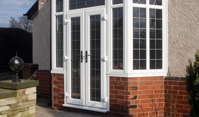 A close-up of a white-framed double French-style door set into a bay window with multiple small rectangular glass panes divided by white mullions in a lead-style grid.