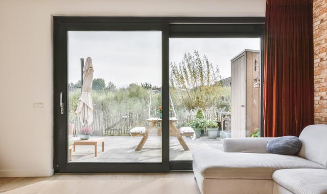 A bright, modern living room with a light-grey sectional sofa next to a large black-framed sliding patio door leading to a small patio. Inside, pale hardwood floors and a red-brown brick accent wall with a deep rust-colored curtain frame the doorway.