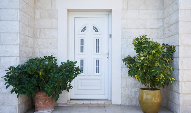 A symmetrical entryway with a white uPVC paneled door set into a light-colored stone wall. The door features decorative raised panels and small vertical glass inserts topped by a shallow arched detail. On either side of the doorway are two potted broadleaf evergreen shrubs.