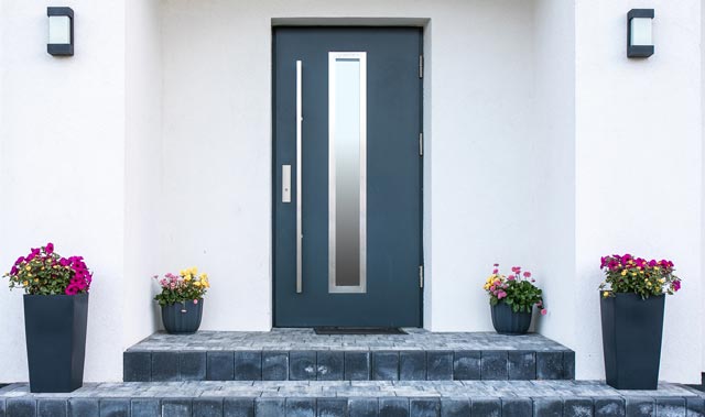 A modern front entryway with a dark-colored door featuring a long vertical metal handle and a narrow vertical glazed panel. Three stone steps lead up to the door, flanked by two tall dark planters with bright flowering plants.
