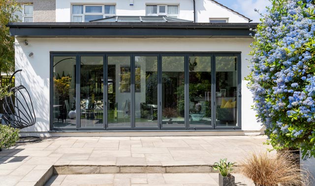 The rear exterior of a modern house featuring a modern extension and wide set of dark-framed glass bi-fold doors with six tall panels, opening onto a paved patio.