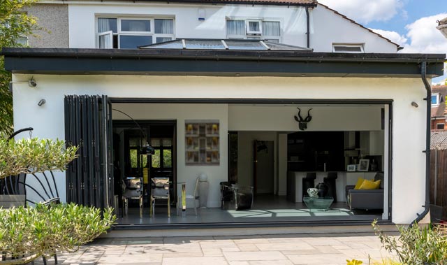 Rear view of a two-story house with a modern single-story brick built orangery featuring large black folding glass doors fully opened onto a paved patio. Inside, a dining area with dark chairs and pendant lights is visible alongside an open-plan living and kitchen space.