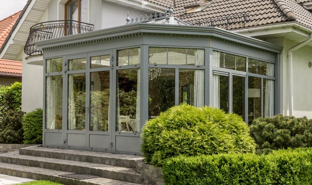 A bright sunroom attached to the back of a house with tall multi-pane glass walls and central glass double doors. The orangery is topped with a decorative roof lantern.