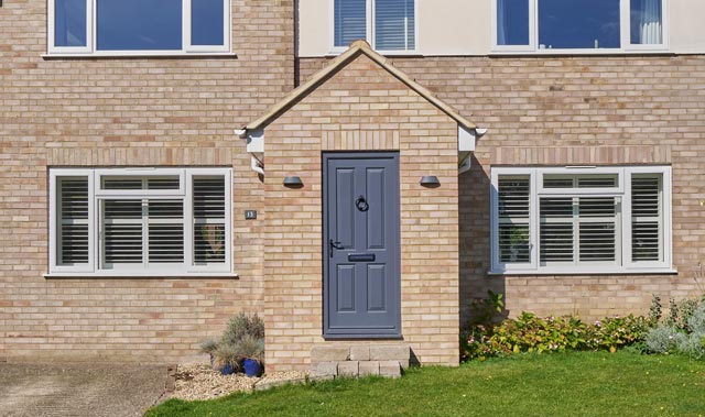 Close-up front view of a modern brick house with light beige bricks and white trim. A small brick built gabled porch with dark blue-gray door featuring a round knocker and mail slot. Two small exterior wall lights flank the door.