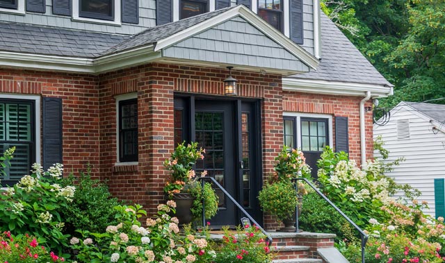 Tidy brick house front with a small enclosed porch featuring a black door with glass sidelights and matching black shutters on windows. Red brick exterior with white trim, gray shingled roof, dormer windows, brick steps with black iron handrails, and lush landscaping.
