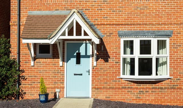 Close-up of a brick house front with a pale blue entrance door under a small gabled open porch with white timber trim and brown tiled roof. The door has a narrow vertical glazed panel, mail slot, and silver handle.