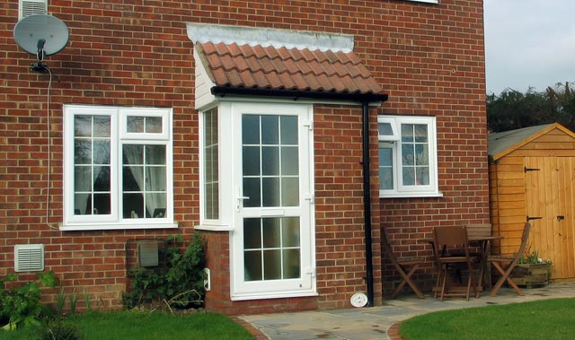 Small brick house exterior with a white-framed uPVC back door under a small tiled canopy porch. White-framed uPVC windows with grid panes flank the door.