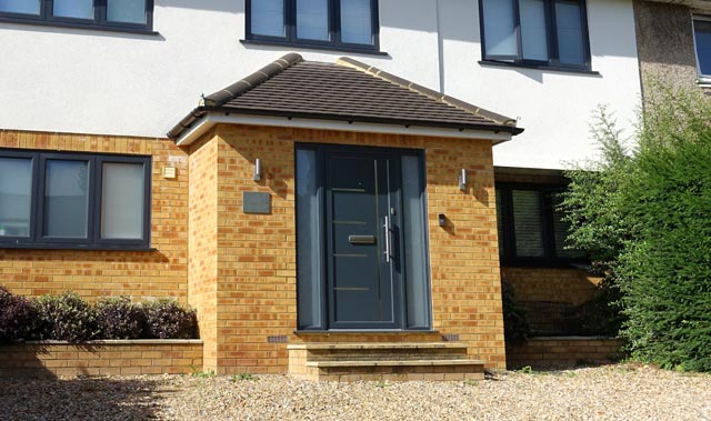 A modern house front entrance with a small brick porch topped by a tiled hipped roof. The dark grey front door has a vertical handle and narrow glazed sidelights. Three steps lead up from a gravel driveway to the porch.