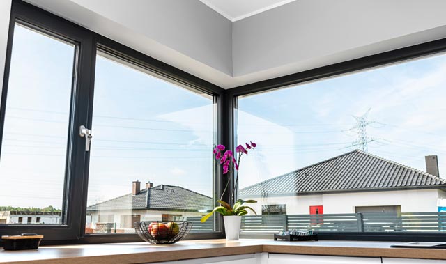 Bright modern kitchen corner with large black-framed corner casement windows overlooking neighboring houses and a clear blue sky.