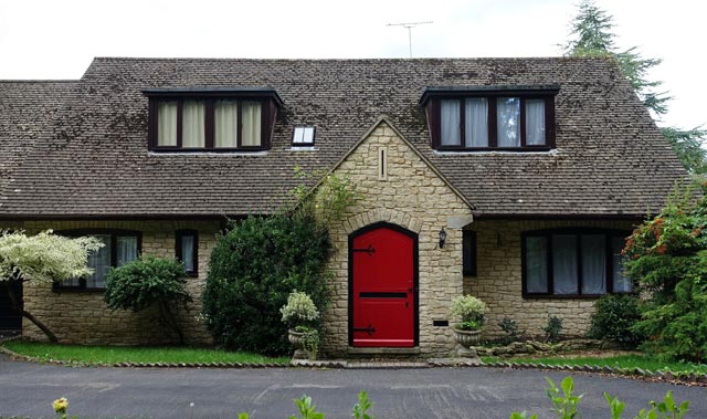 Cottage-style house with steep dark-shingled roof, light tan stone exterior, and bright red arched wooden front door. The house features dark-framed dormer and ground floor cottage windows with curtains, surrounded by shrubs, urn planters, and a paved driveway.