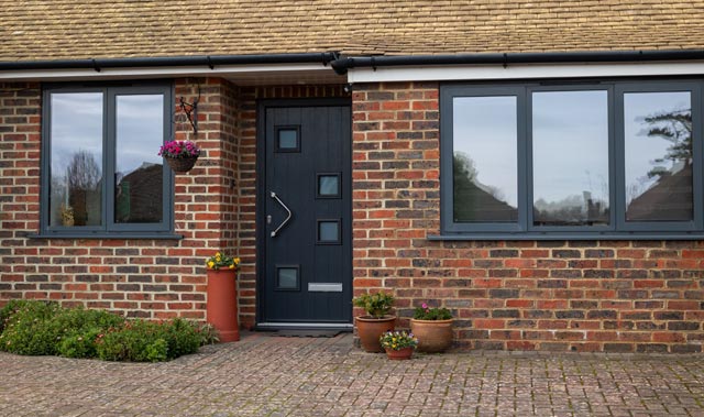 Front view of a tidy single-story brick house with dark charcoal front door. The house has dark-framed flush casement windows, a light tan tiled roof, black guttering, a paved driveway, and well-kept landscaping.