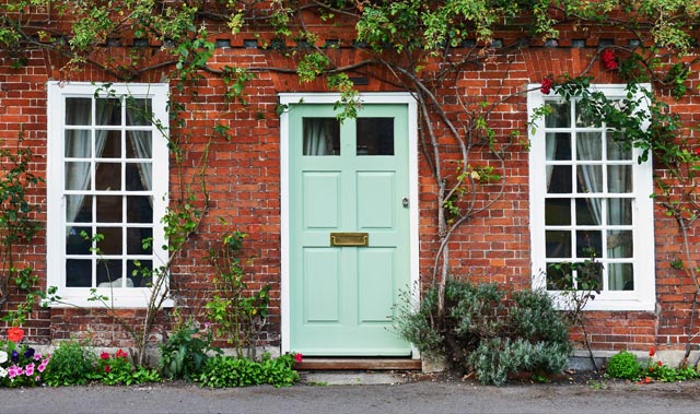 Charming brick-front cottage with pale seafoam-green wooden door featuring a small glazed panel and brass mail slot. Tall white-framed multi-pane sash windows flank the door, surrounded by climbing vines with red blooms.