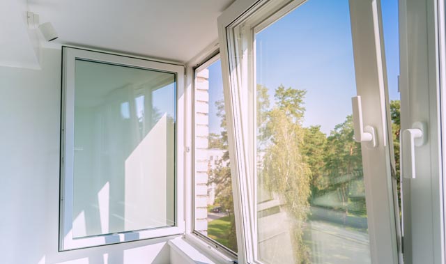 Interior corner of a bright modern room with large white-framed tilt and turn windows; one window fully open inward and the other tilted for ventilation.