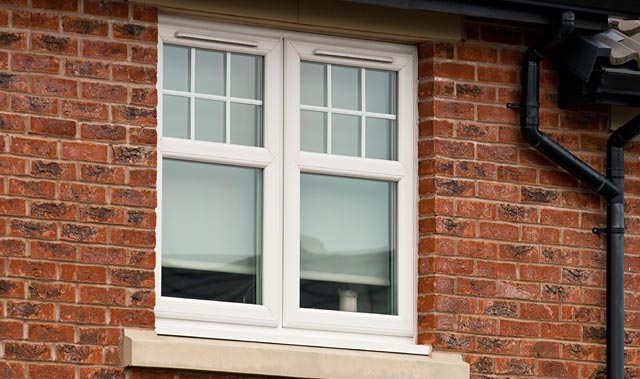 White-framed uPVC double window with decorative upper panes set in a red brick wall, featuring a light stone sill and a black rain gutter on the side.
